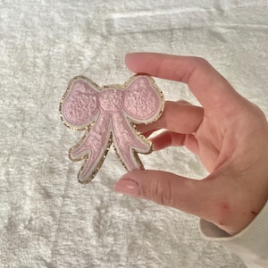 Puede incluir: Un lazo rosa y dorado con purpurina y un contorno blanco. El lazo es un parche de tela.