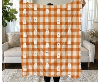 May include: Orange and white plaid blanket featuring repeating white ghost illustrations. The blanket is held up, showcasing the pattern. The design is reminiscent of Halloween decor, with a cozy and festive feel.