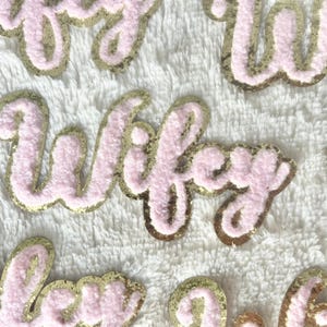 Peut inclure: Patchs en feutre rose et or avec le mot "wifey" écrit dessus.
