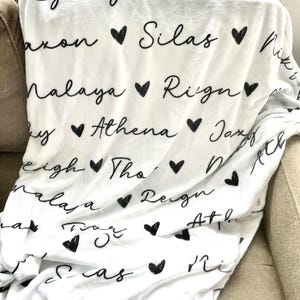 May include: White fleece blanket with black handwritten names and hearts. The names include Rylergh, Thar, axon, Silas, Niks, Nalaya, Rign, by, Athena, Jaxy, ch, alaa, Reign, Ath, Atl, Sas, Ri, Riegn.