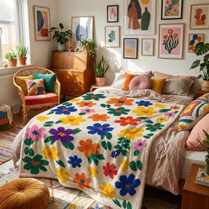 Puede incluir: Un dormitorio con una cama cubierta por una manta crema adornada con grandes y coloridas flores en tonos azules, amarillos, naranjas, verdes y morados. La habitación está decorada con arte enmarcado, plantas y una silla de mimbre con cojines coloridos.