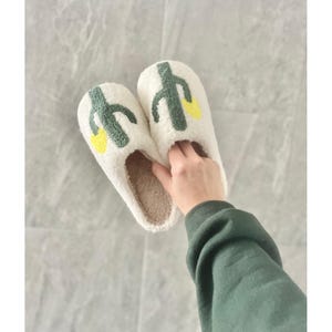 May include: A pair of off-white, plush slippers with a cactus design in green and yellow. The slippers have a soft, brown interior and are designed for indoor wear. The image shows the slippers being held.
