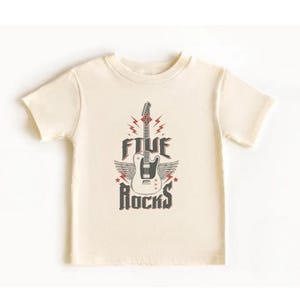 May include: A cream colored short-sleeved t-shirt with a graphic of a white electric guitar with wings and the text "FIVE ROCKS" in black and red.