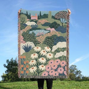 May include: A woven throw blanket featuring a colorful landscape scene with a small house, a pond, and lush garden foliage. The design includes white daisies, pink flowers, and green shrubbery with a fringed edge on the top and bottom sides.