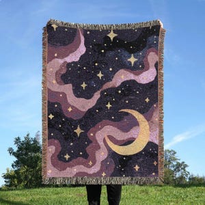 Könnte beinhalten: Eine gewebte Wohndecke mit dunkelblauem Hintergrund, wirbelnden violetten und rosafarbenen Nebelwolken, einem großen goldgelben Halbmond und verstreuten vierzackigen Sternen. Die Decke hat an der Ober- und Unterseite einen Fransenrand.