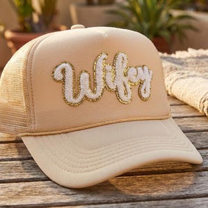 May include: A beige trucker hat with a mesh back and a curved brim. The word "Wifey" is embroidered on the front in white script with gold glitter accents. The hat is sitting on a wooden surface.
