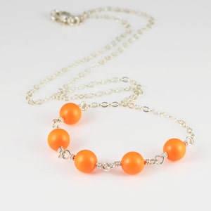 Könnte beinhalten: Eine silberne Kettenkette mit fünf leuchtend orangefarbenen Perlen. Die Perlen sind gleichmäßig verteilt und mit Silberdraht verbunden.