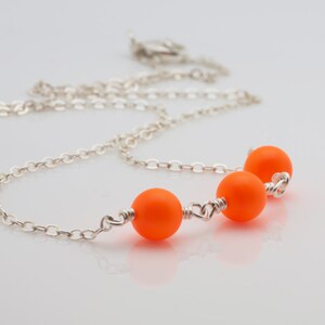 Op de afbeelding: Een zilveren ketting met drie feloranje kralen. De kralen zijn met zilverdraad aan de ketting bevestigd.