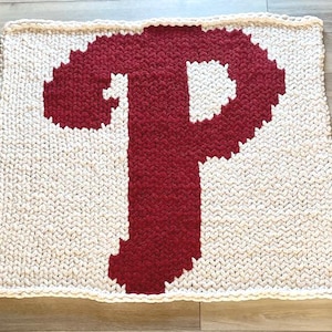 May include: A square-shaped chunky knit blanket featuring a large, bold letter P in deep red yarn centered against a cream-colored background. The textile is crafted with thick, interlocking loops of yarn, creating a textured, soft appearance.