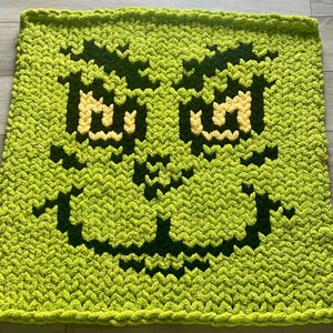 Könnte beinhalten: Eine hellgrüne Häkeldecke mit einem pixeligen Bild eines Gesichts mit gelben Augen und einer dunkelgrünen Umrandung. Die Decke ist quadratisch und hat ein strukturiertes Aussehen.