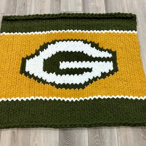 May include: Hand-knitted blanket in green, gold, and white. The blanket features a large white letter "G" on a gold background, with green borders and white stripes. The blanket is made of thick, chunky yarn.