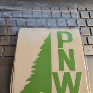 May include: A stack of white vinyl stickers featuring a green evergreen tree silhouette and the letters "PNW" in green. The stickers are in a clear plastic bag, resting on a laptop keyboard.