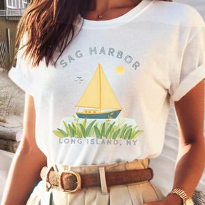 Peut inclure: T-shirt blanc avec un motif graphique représentant un voilier sur l'eau avec le texte "Sag Harbor Long Island, NY".