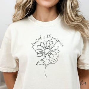 May include: Cream-colored t-shirt featuring a black line drawing of a daisy with the words "planted with purpose" in a script font. The shirt has short sleeves and a crew neck.