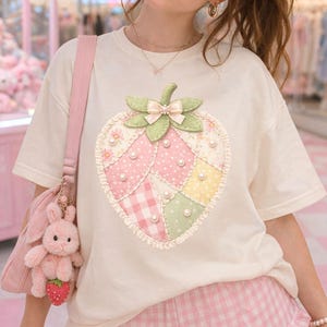 May include: Cream-colored t-shirt featuring a large, patchwork strawberry design with pink, green, and yellow sections, adorned with pearls. The strawberry has a green stem and bow. The model is wearing pink heart-shaped sunglasses and pink gingham shorts.