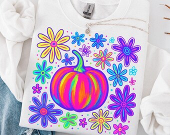 May include: White sweatshirt featuring a vibrant, colorful pumpkin and flower design. The pumpkin is a mix of pink, orange, and yellow, surrounded by blue, purple, and yellow flowers. A silver necklace rests on the shirt.