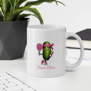 Peut inclure: Une tasse à café en céramique blanche avec un cornichon de dessin animé vert portant un bandeau rose et tenant une pagaie rose. Le cornichon porte des baskets roses et le texte "Dink Diva" est imprimé sous le cornichon.
