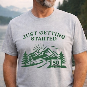 以下が含まれることがあります： 「JUST GETTING STARTED」の文字と、道路と数字の50が入った緑色の山のグラフィックがプリントされた、ライトグレーのTシャツ。半袖です。
