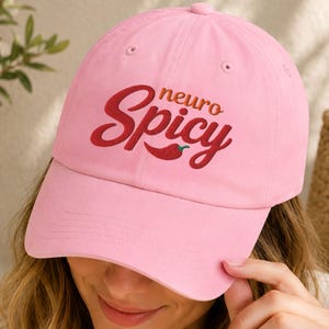 Op de afbeelding: Een lichtroze baseballpet met de geborduurde tekst "neuro Spicy" in een donkerrood cursief lettertype, vergezeld van een klein rood chilipeper-icoontje op het voorpaneel. De pet heeft een gebogen klep en een gestructureerd ontwerp.