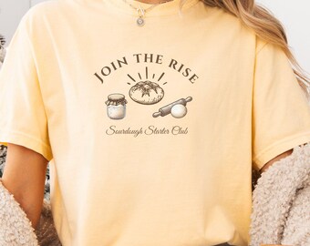 Könnte beinhalten: Hellgelbes T-Shirt mit dem Text "JOIN THE RISE" über einer Illustration von einem Glas, Brot und einem Nudelholz. Darunter steht der Text "Sourdough Starter Club". Das Shirt hat einen lässigen Stil.