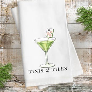 Puede incluir: Toalla de cocina blanca con una ilustraci&oacute;n de c&oacute;ctel verde en una copa de Martini. La copa tiene una etiqueta decorativa con caracteres japoneses y flores. El texto "TINIS & TILES" est&aacute; impreso en negro debajo.