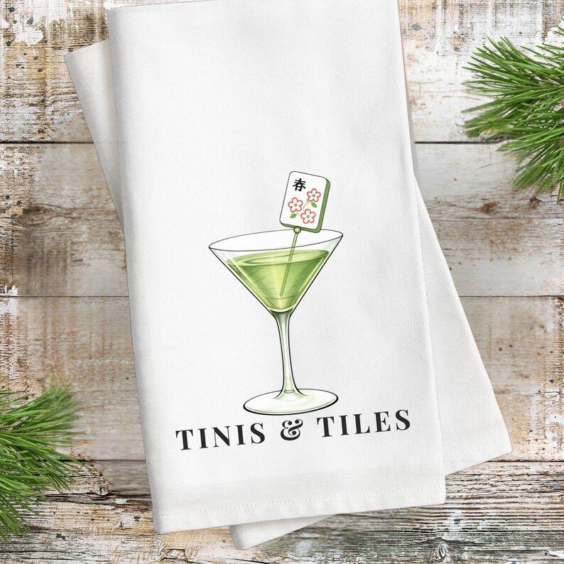 Puede incluir: Toalla de cocina blanca con una ilustraci&oacute;n de c&oacute;ctel verde en una copa de Martini. La copa tiene una etiqueta decorativa con caracteres japoneses y flores. El texto "TINIS & TILES" est&aacute; impreso en negro debajo.