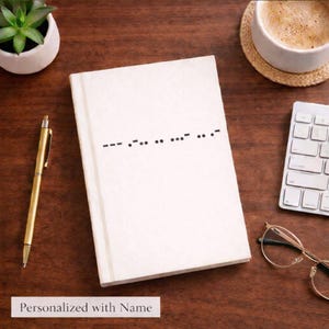 May include: A white notebook with a Morse code pattern on the cover, resting on a wooden surface. A gold pen, a small plant, a cup of coffee, glasses, and a keyboard are also on the table. The text "Personalized with Name" is at the bottom.
