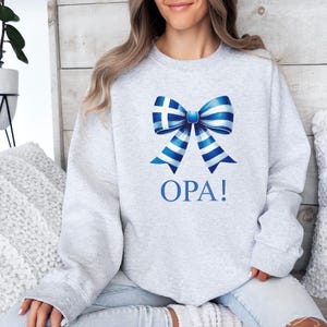 May include: A gray sweatshirt with a blue and white bow featuring the Greek flag and the word "OPA!" printed on the front.