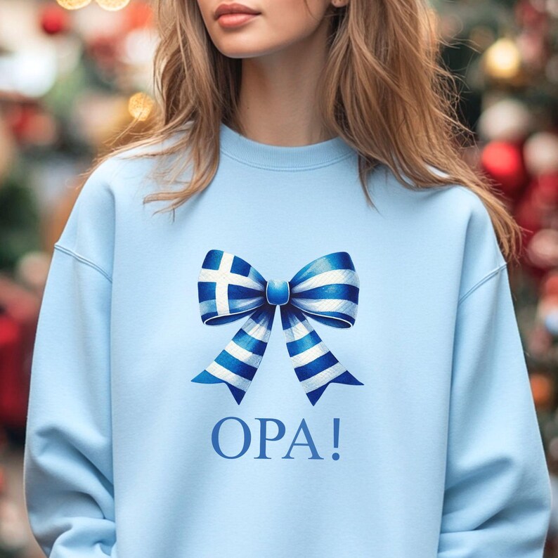 May include: A light blue sweatshirt with a white and blue bow printed on the front. The bow is designed with the Greek flag colors and pattern. The text "OPA!" is printed below the bow.