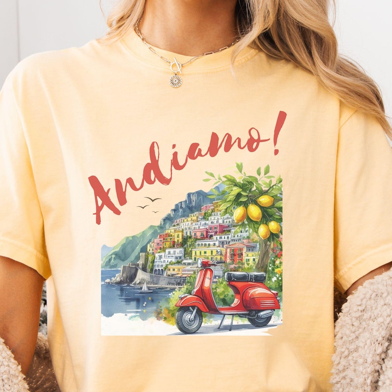 K&ouml;nnte beinhalten: Ein gelbes T-Shirt mit einer bunten Illustration eines Rollers, einer Stadtlandschaft und eines Zitronenbaums. Der Text "Andiamo!" ist in Rot auf dem Shirt gedruckt.