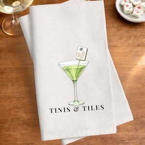 May include: White kitchen towel with a martini glass illustration and the text "TINIS & TILES". The martini glass contains a green liquid and a decorative garnish. Mahjong tiles are scattered around the towel.