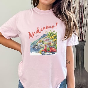 K&ouml;nnte beinhalten: Rosafarbenes T-Shirt mit einem Motiv eines roten Rollers, der durch ein farbenfrohes italienisches Dorf f&auml;hrt, mit dem Text "Andiamo!" auf der Vorderseite.
