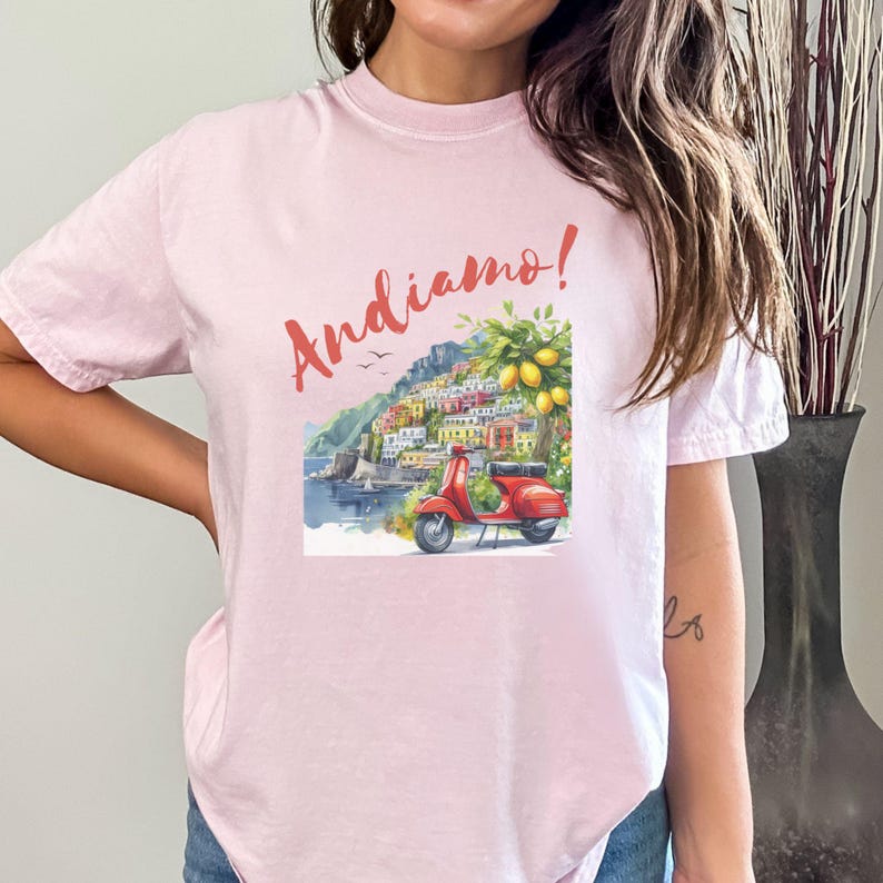 K&ouml;nnte beinhalten: Rosafarbenes T-Shirt mit einem Motiv eines roten Rollers, der durch ein farbenfrohes italienisches Dorf f&auml;hrt, mit dem Text "Andiamo!" auf der Vorderseite.
