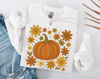 May include: White sweatshirt featuring a large orange pumpkin with gold glitter accents, surrounded by orange, yellow, and green flowers. A silver necklace with a small pendant rests on the shirt. The sweatshirt is on a white surface with a pair of blue jeans.
