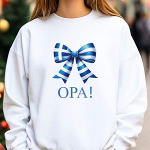 May include: White sweatshirt with a blue and white bow printed on the front. The bow is designed with the Greek flag colors and pattern. The text "OPA!" is printed below the bow.