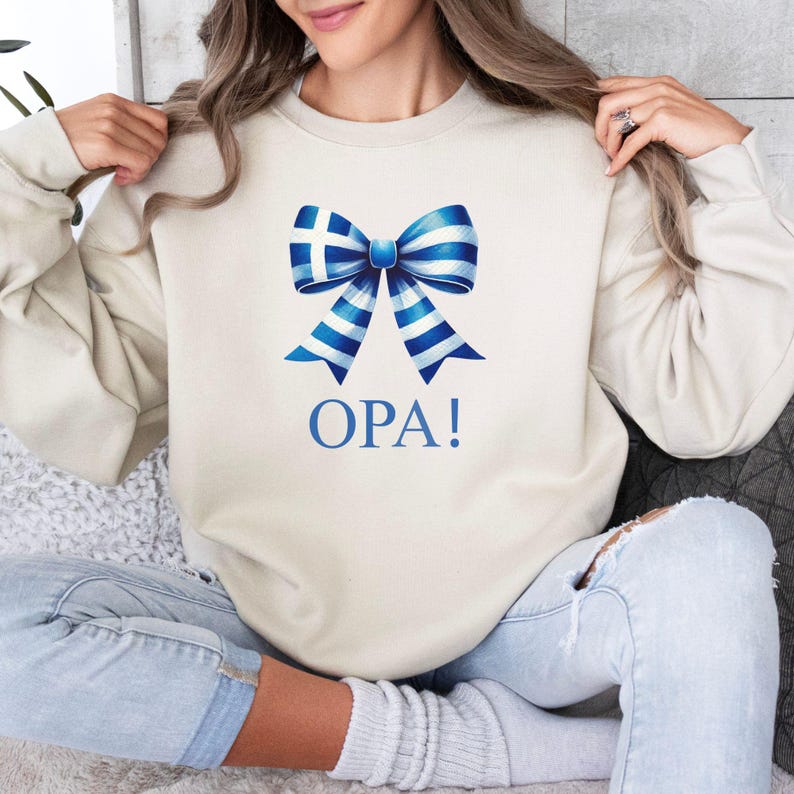 May include: A cream-colored sweatshirt with a blue and white bow with a Greek flag design. The text "OPA!" is printed below the bow.