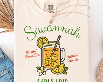 Può includere: T-shirt color crema con la scritta "Savannah" in verde. Sotto, una grafica di un barattolo con tè freddo, fette di limone e una cannuccia. Il testo include "Sippin' Sweet Tea", "Spillin' Secrets" e "GIRLS TRIP 2025".
