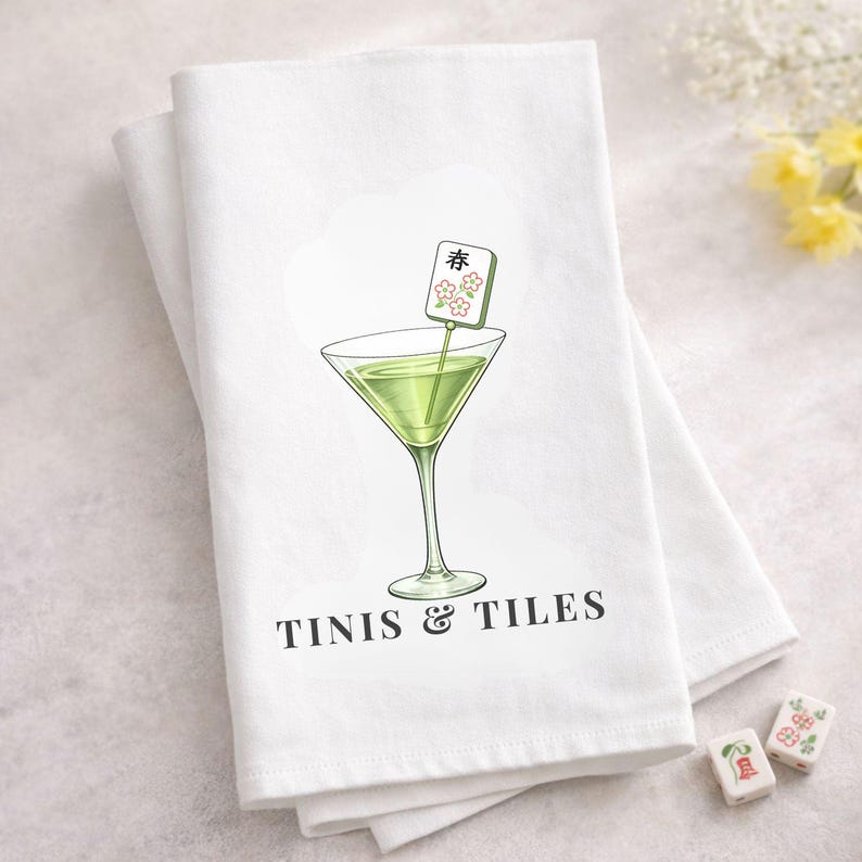 May include: White kitchen towel with a martini glass illustration and the text "TINIS & TILES". The martini glass contains a green liquid and a decorative garnish. Mahjong tiles are scattered around the towel.