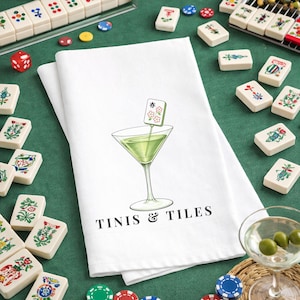 Puede incluir: Toalla de cocina de algod&oacute;n blanco con una ilustraci&oacute;n de c&oacute;ctel verde y el texto "Tinis & Tiles". El c&oacute;ctel tiene una guarnici&oacute;n de ficha de Mahjong. Fichas de Mahjong, dados y fichas rodean la toalla sobre una superficie verde.