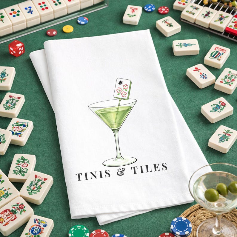 Puede incluir: Toalla de cocina de algod&oacute;n blanco con una ilustraci&oacute;n de c&oacute;ctel verde y el texto "Tinis & Tiles". El c&oacute;ctel tiene una guarnici&oacute;n de ficha de Mahjong. Fichas de Mahjong, dados y fichas rodean la toalla sobre una superficie verde.