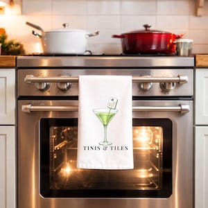 May include: White kitchen towel with a martini glass illustration and the text "TINIS & TILES". The martini glass contains a green liquid and a decorative garnish. Mahjong tiles are scattered around the towel.