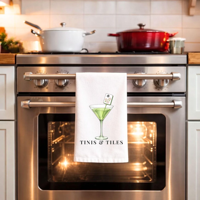 May include: White kitchen towel with a martini glass illustration and the text "TINIS & TILES". The martini glass contains a green liquid and a decorative garnish. Mahjong tiles are scattered around the towel.
