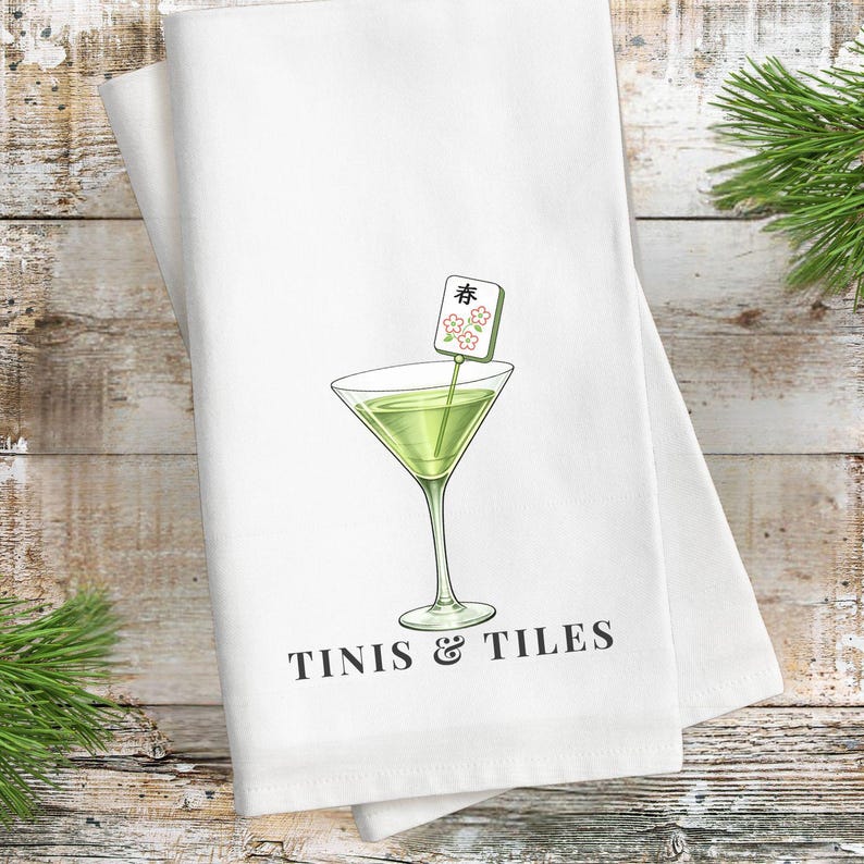 May include: White kitchen towel with a martini glass illustration and the text "TINIS & TILES". The martini glass contains a green liquid and a decorative garnish. Mahjong tiles are scattered around the towel.