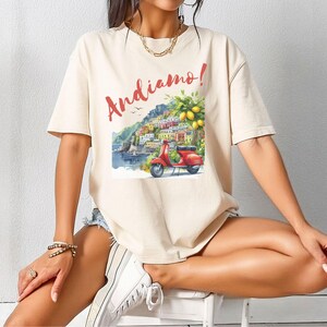 K&ouml;nnte beinhalten: Ein cremefarbenes T-Shirt mit einem grafischen Druck eines roten Rollers, der durch ein farbenfrohes italienisches Dorf f&auml;hrt, mit dem Text "Andiamo!" in Rot.