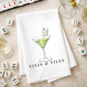 Puede incluir: Pa&ntilde;o de cocina blanco con una ilustraci&oacute;n de copa de Martini y el texto "TINIS & TILES". La copa de Martini contiene un l&iacute;quido verde y una guarnici&oacute;n. Azulejos de Mahjong esparcidos.