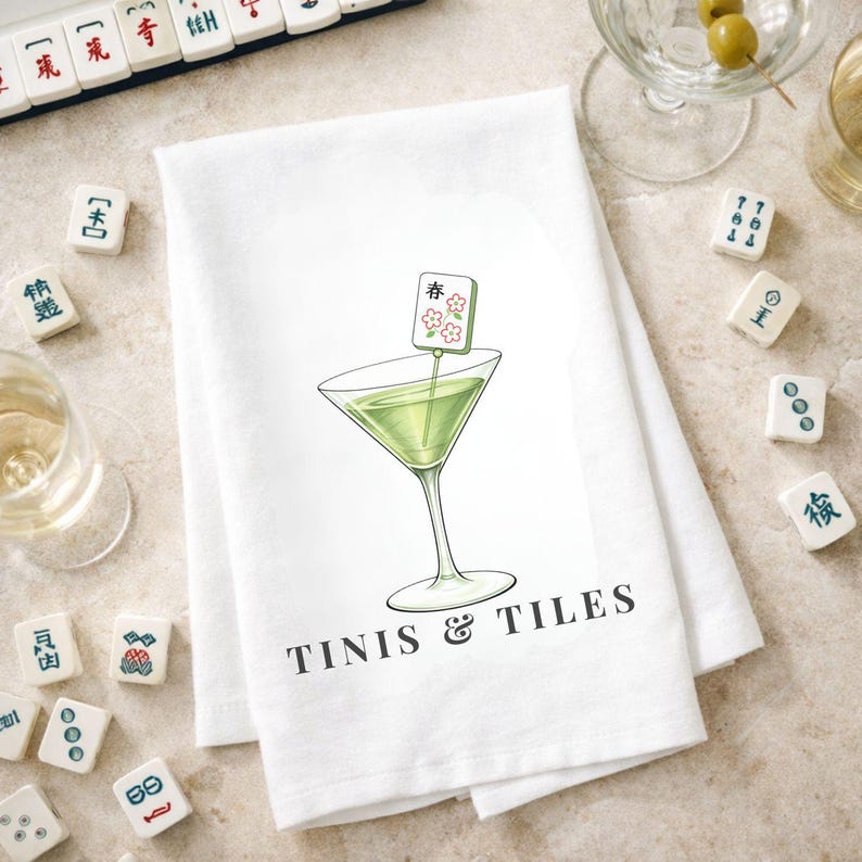 Puede incluir: Pa&ntilde;o de cocina blanco con una ilustraci&oacute;n de copa de Martini y el texto "TINIS & TILES". La copa de Martini contiene un l&iacute;quido verde y una guarnici&oacute;n. Azulejos de Mahjong esparcidos.