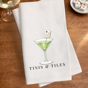 Puede incluir: Toalla de cocina blanca con un dise&ntilde;o de copa de martini verde y una ficha de Mahjong. La toalla tiene las palabras "Tinis & Tiles" impresas en negro. El dise&ntilde;o est&aacute; centrado en la toalla.