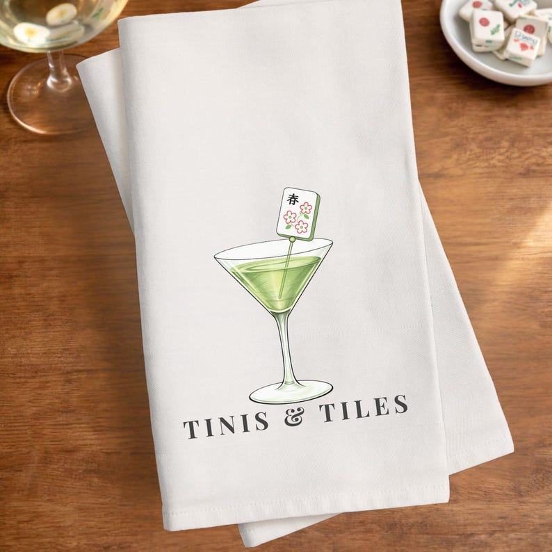 Puede incluir: Toalla de cocina blanca con un dise&ntilde;o de copa de martini verde y una ficha de Mahjong. La toalla tiene las palabras "Tinis & Tiles" impresas en negro. El dise&ntilde;o est&aacute; centrado en la toalla.