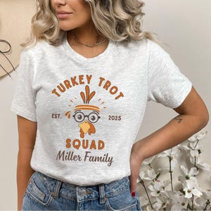 May include: A light gray t-shirt with the text "TURKEY TROT SQUAD Miller Family" in brown. The graphic features a cartoon turkey wearing glasses and a headband. The shirt also has the text "EST. 2025".