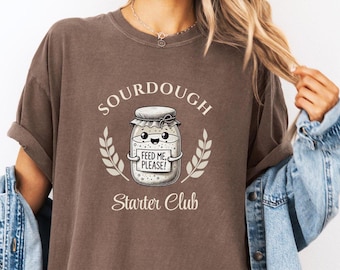 May include: A brown t-shirt with a sourdough starter jar graphic. The jar has a smiling face and a sign that says "Feed me, please!" The text "Sourdough Starter Club" is below the jar.
